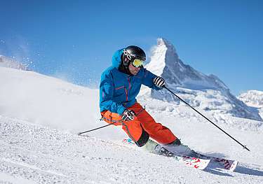 Skifahrer in blauer Jacke und orangefarbener Hose fährt vor verschneiten Bergen.