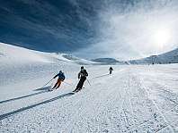 Trois skieurs descendent une large piste enneigée et ensoleillée en montagne.