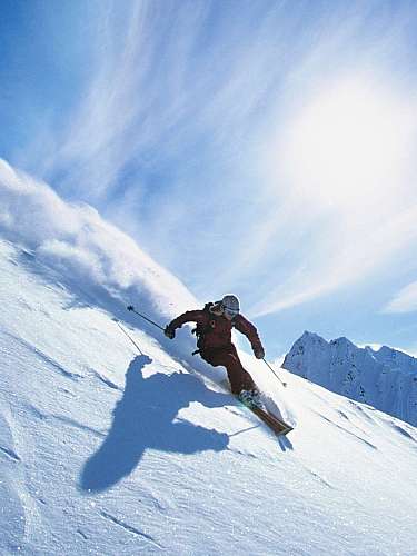 Ein Skifahrer fährt eine steile, verschneite Bergflanke unter blauem Himmel hinab.
