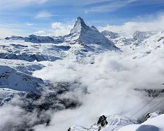 Paysage montagneux enneigé avec sommet, nuages et station de téléphérique au premier plan.